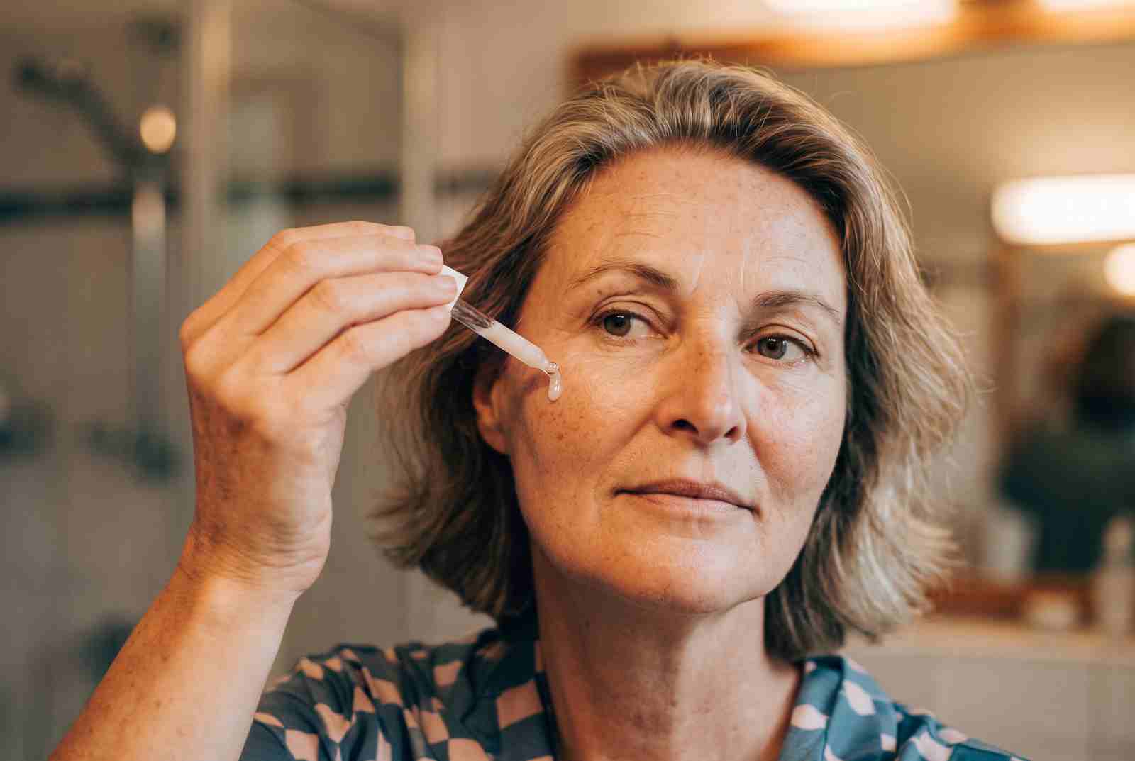 Marion trägt am Badezimmerspiegel mit einer Pipette Serum auf ihre Wange auf