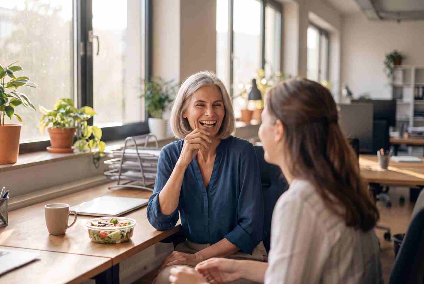 Birgit lacht mit einer jüngeren dunkelhaarigen Kollegin im hellen Büro, Salat und Kaffeetasse auf dem Schreibtisch