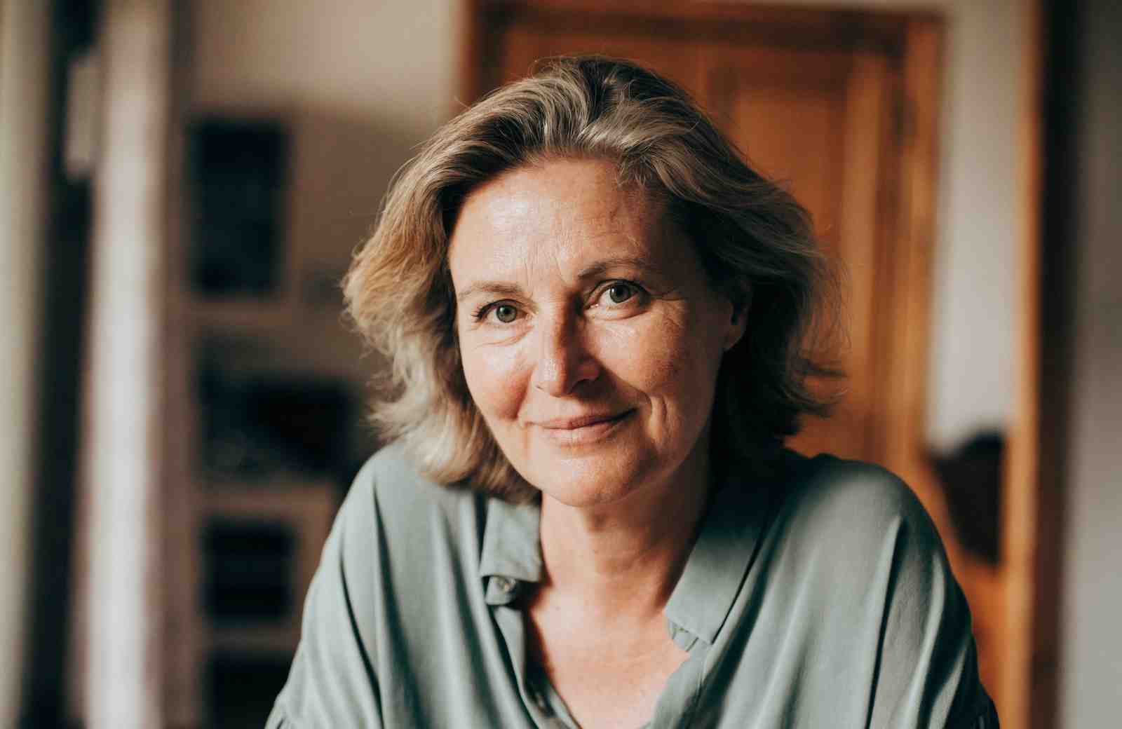 Marion (53) mit blond-grauem, welligem Haar in salbeigrüner Bluse, warmes Licht, Blick in die Kamera